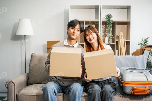 Preview: A young couple happily sits on sofa, packing their belongings into a big box for their move to