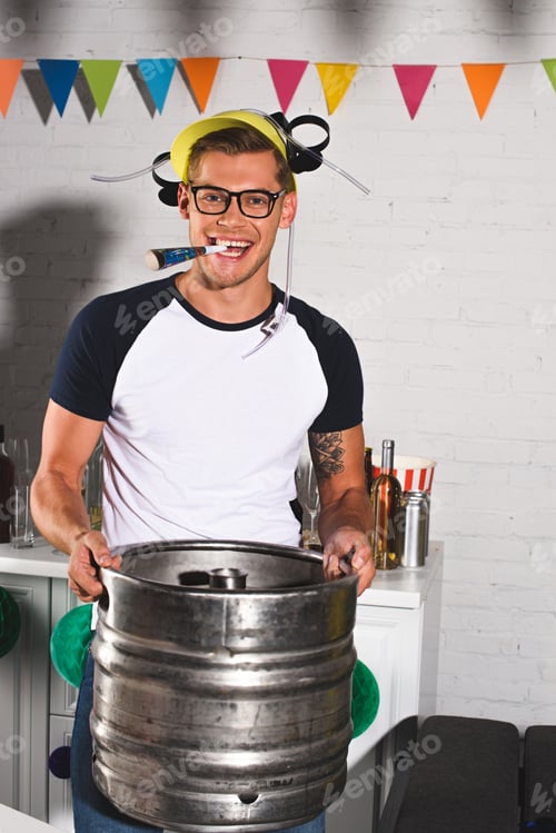 Preview: happy young man in beer hat holding keg of beer and smiling at camera