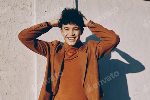 Preview: Young man with curly hair smiling joyfully, hands on head, against a textured light wall Warm autumn
