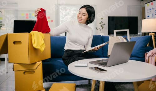 Preview: Asian female entrepreneur checking clothes in cardboard box and taking notes at home