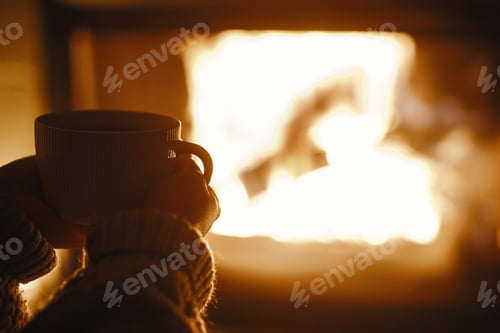 Preview: Woman holding cup of tea and warming up hands at cozy fireplace in dark evening room