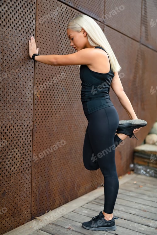 Preview: Side view of fit woman stretching after workout against metal wall in the city