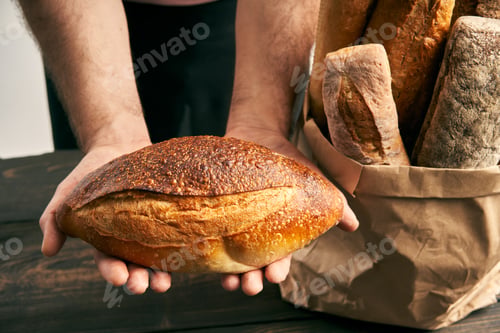 Preview: Baker man in apron holding loaf of fresh baked bread in the hands