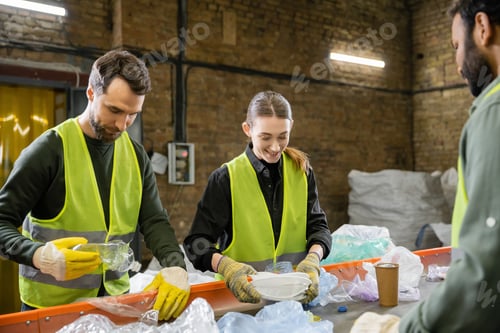 Preview: Positive young worker in gloves and protective clothes sorting garbage on conveyor near multiethnic