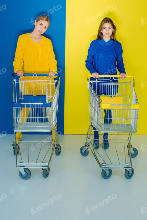 Preview: Pretty girls in fashion clothes pushing shopping carts on blue and yellow background