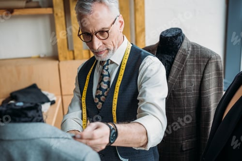 Preview: handsome mature tailor with measuring tape looking at jacket on mannequin at sewing workshop