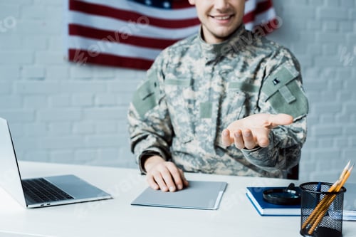 Preview: cropped view of happy soldier gesturing near laptop in office