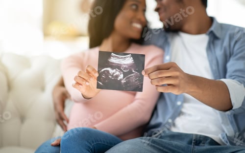 Preview: Positive black couple showing ultrasound photo of their unborn baby at home, selective focus on