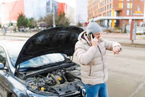 Preview: A forlorn man stands beside his broken car, with a troubled expression as he speaks on his phone,