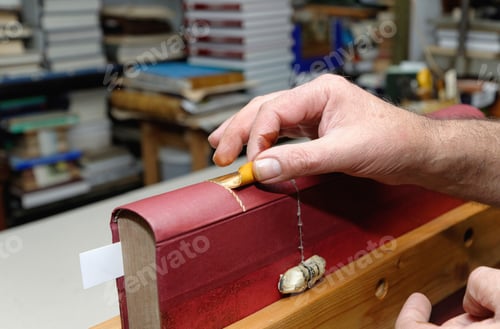 Preview: Close up of hands of senior male traditional bookbinder applying gold leaf to book spine
