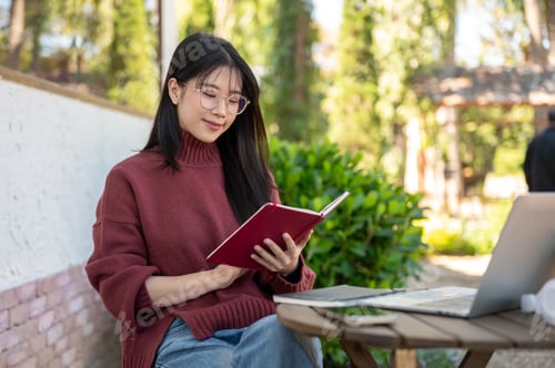 Preview: Glasses asian woman in sweater reading book aside laptop and sitting at wooden table in garden cafe.