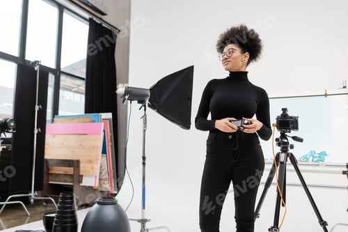 Preview: african american content producer with exposure meter looking away near softbox reflector and