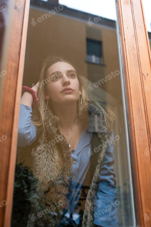Preview: Young woman lost in thought, gazing through a window pane.