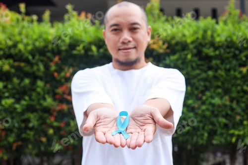 Preview: Smiling Asian young man hands showing light blue ribbon awareness on palms.