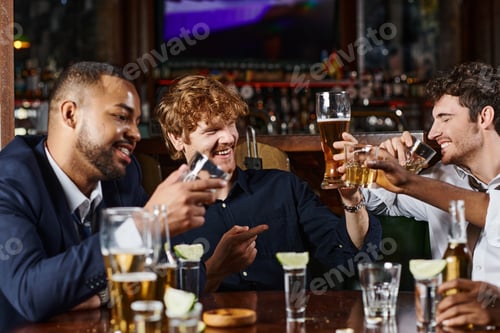Preview: happy redhead man pointing at friend while having drinks after work in bar, interracial friends