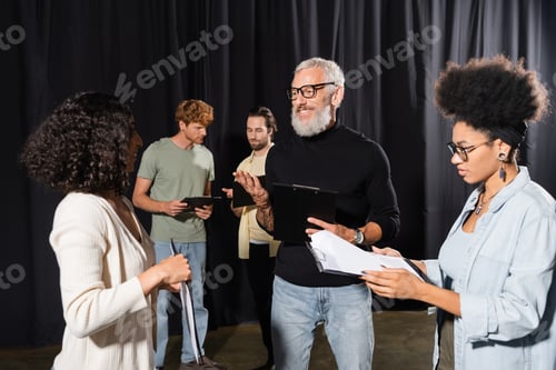 Preview: bearded screenwriter talking to interracial actresses holding screenplays in theater school.