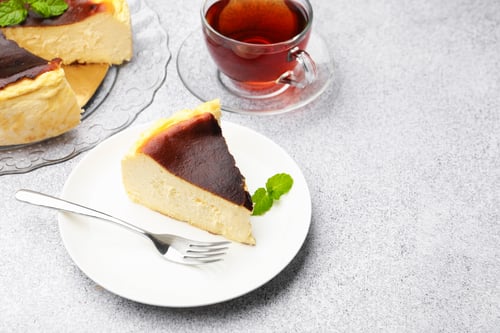 Preview: Piece of delicious Basque cheesecake with mint and tea on light grey table, closeup. Space for text