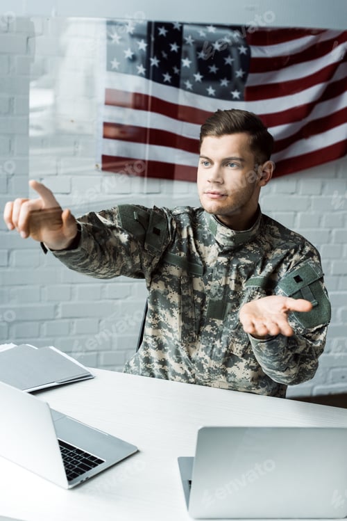 Preview: handsome soldier in uniform gesturing near laptops in office