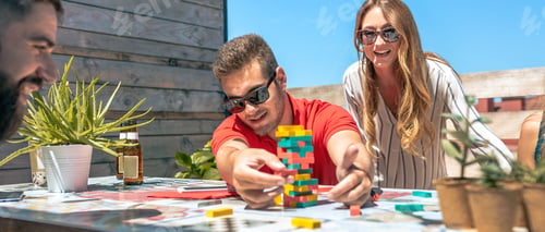 Preview: Concentrated man catching jenga game piece next to colleagues on terrace