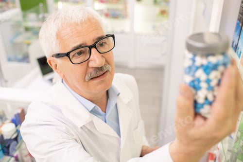 Preview: Aged drugstore worker in whitecoat and eyeglasses looking at big bottle of pills