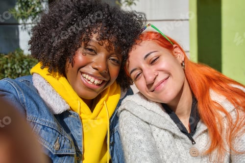Preview: two young friends smiling taking a selfie one black afro hair and the other white orange hair