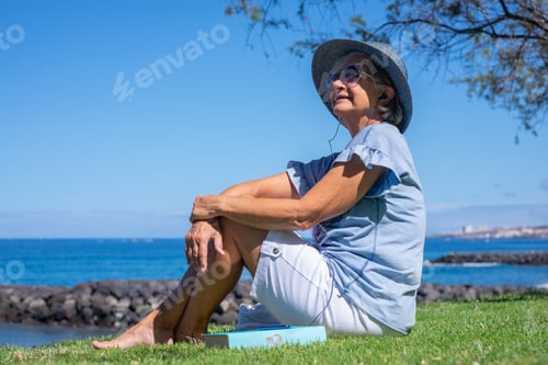 Preview: Smiling senior woman in hat sitting barefoot in meadow in front of sea listening to music, relaxed s