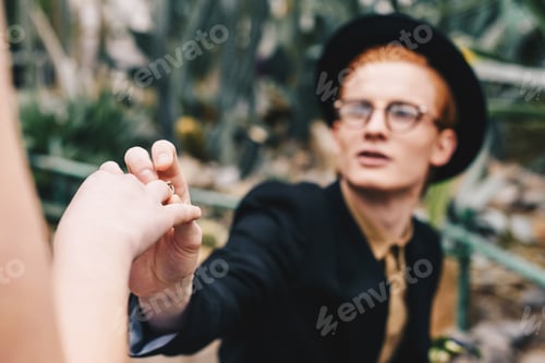 Preview: cropped shot of stylish young man in hat and eyeglasses making proposal and putting ring to girl