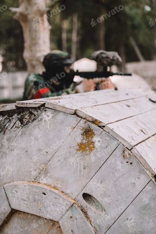 Preview: selective focus of paintball splashes on wooden barrel and paintball player shooting behind outdoors