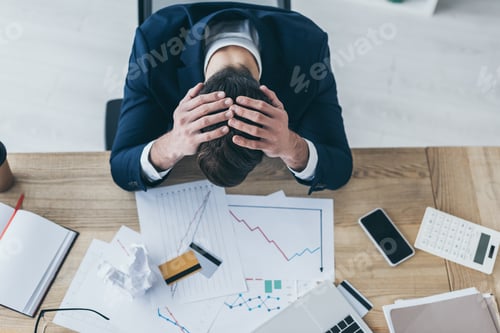 Preview: top view of depressed businessman holding hands on bowed head while sitting at desk with documents