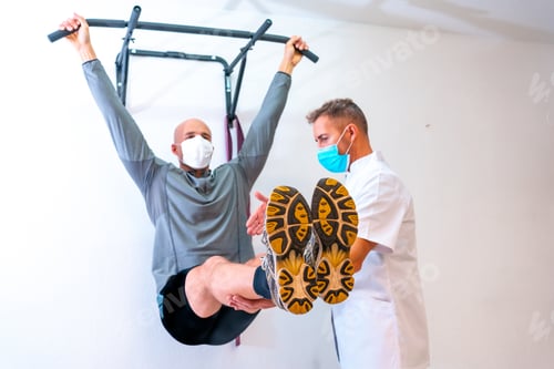 Preview: Physiotherapist with mask stretching client's back and arms against wall.