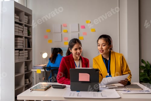 Preview: Two women are sitting at a desk with a laptop and papers in front of them