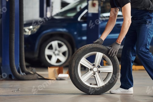 Preview: Close up view of man in work uniform with car wheel indoors. Conception of automobile service