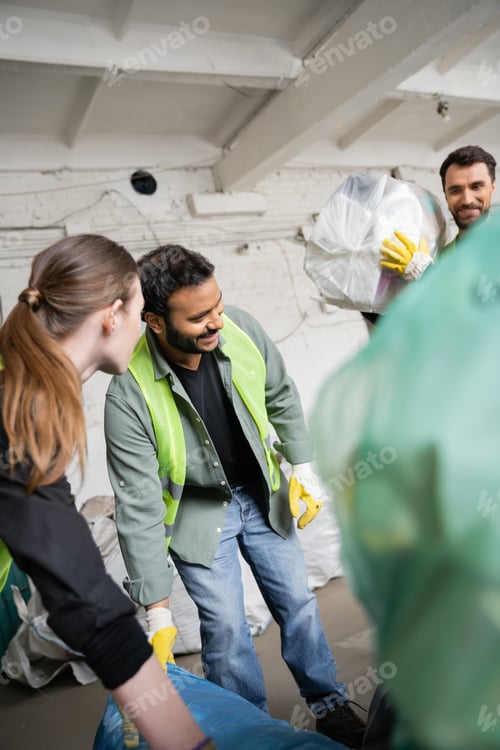 Preview: Smiling indian worker in high visibility vest and gloves working near colleagues and plastic bags in