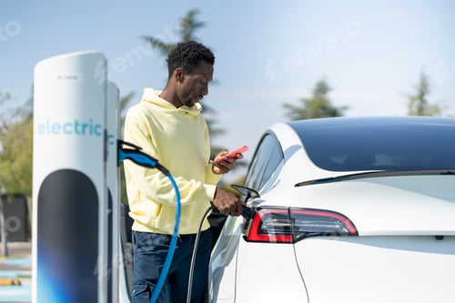 Preview: African american man charging modern electric car at public station while using phone