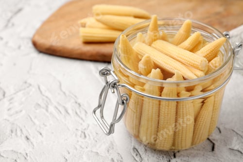 Preview: Jar of pickled baby corn on white textured table, closeup. Space for text