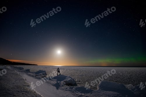 Preview: Traveler on the seashore covered with ice under a starry sky with moon and northern lights.