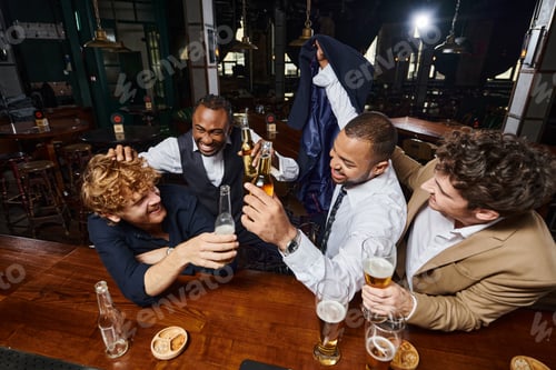 Preview: group of four happy colleagues in formal wear joking and drinking beer in bar, having fun after work