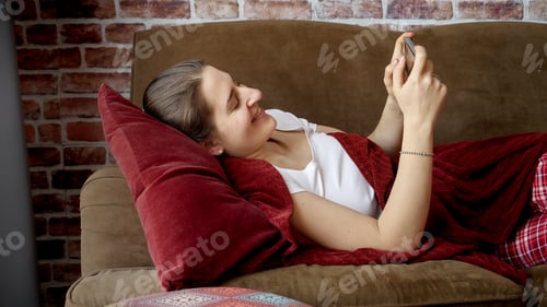 Preview: Young smiling woman resting on couch in living room and using smartphone