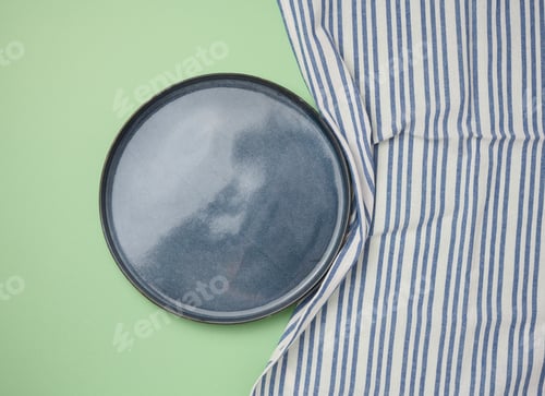 Preview: Empty round ceramic plate and blue kitchen towel on green background, top view
