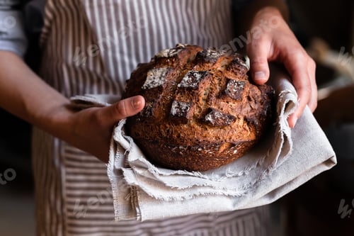Preview: Young woman holding freshly baked dark round bread in her hands.
