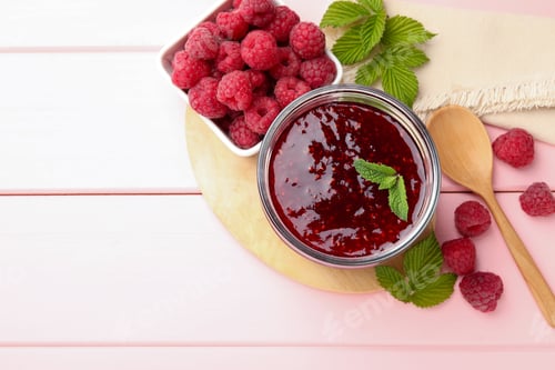 Vista previa: Mermelada dulce de frambuesa en tarro de cristal, bayas y menta sobre mesa de madera rosa, puesta en posición plana. Espacio para texto