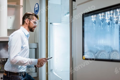 Preview: Businessman with tablet wearing safety goggles examining machine in factory looking