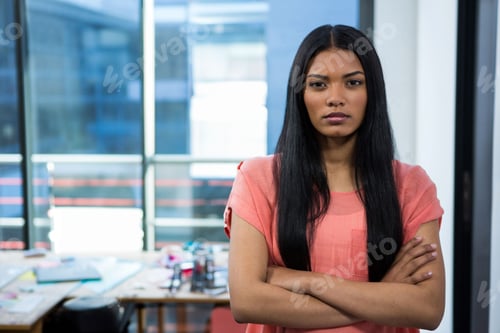 Preview: Standing woman crossing arms in design studio by windows, with cluttered desk and supplies