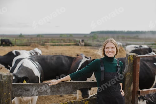 Preview: Smiling Female Farmer in Overalls Standing by Fence with Cows in the Background at a Rural Farm