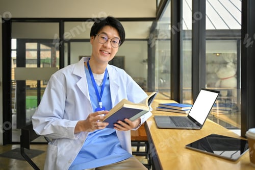 Preview: Doctor in glasses and white coat reading book and using laptop in hospital canteen.