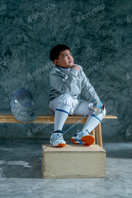 Preview: Vertical image of boy wear fencing sport uniform also hold epee and sit on wooden bench.