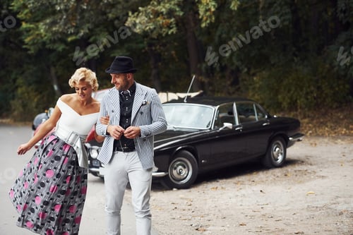 Preview: Beautiful couple in old fashioned wear near retro car with forest at background