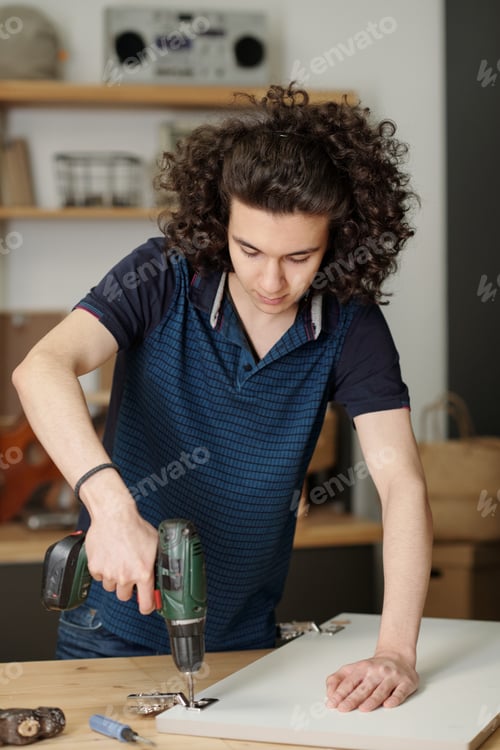 Preview: Youthful guy with dark wavy hair drilling plywood door of kitchen cabinet