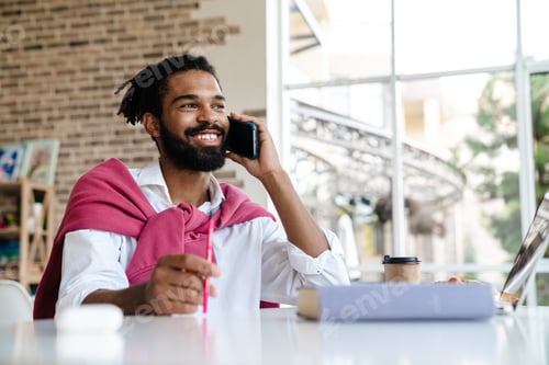 Preview: Smiling african american guy talking on cellphone and working with laptop