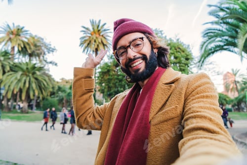 Preview: Bearded young caucasian male taking a selfie portrait with a smartphone in a park outdoors, wearing